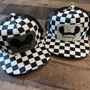 K&ouml;nnte beinhalten: Zwei schwarz-wei&szlig; karierte Trucker-Caps. Eine Kappe hat einen grauen Aufn&auml;her mit einer Mickey Mouse-Silhouette und dem Wort "Daddy". Die andere Kappe hat eine Minnie Mouse-Silhouette und das Wort "mommy".