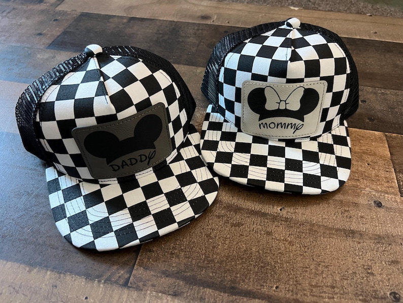 K&ouml;nnte beinhalten: Zwei schwarz-wei&szlig; karierte Trucker-Caps. Eine Kappe hat einen grauen Aufn&auml;her mit einer Mickey Mouse-Silhouette und dem Wort "Daddy". Die andere Kappe hat eine Minnie Mouse-Silhouette und das Wort "mommy".