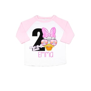 May include: A white and pink baseball tee shirt with a black number 2 and a cartoon duck wearing sunglasses. The text "Best Day Ever" and the name "Emma" are printed on the shirt.