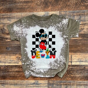 May include: A green, bleach-splattered t-shirt with a cartoon image of Mickey Mouse wearing sunglasses and a checkered background. The name "DEVIN" is spelled out in colorful letters below the image.