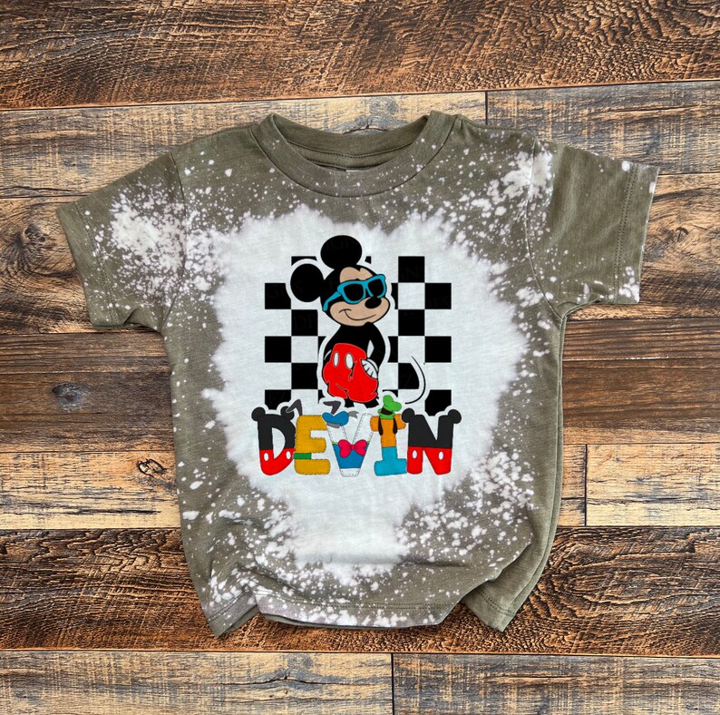 May include: A green, bleach-splattered t-shirt with a cartoon image of Mickey Mouse wearing sunglasses and a checkered background. The name "DEVIN" is spelled out in colorful letters below the image.