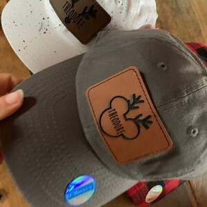 May include: Two baseball caps with brown leather patches featuring a reindeer design and the text "Theodore". One cap is white with black speckles and the other is gray.