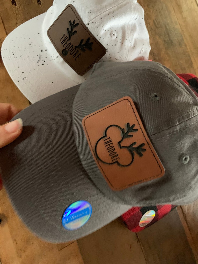 May include: Two baseball caps with brown leather patches featuring a reindeer design and the text "Theodore". One cap is white with black speckles and the other is gray.