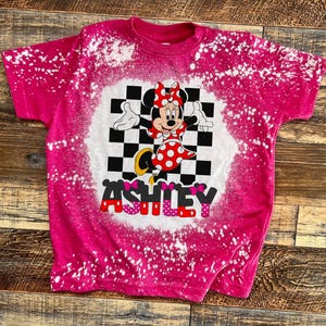 May include: Pink t-shirt with a bleach-splattered design featuring a cartoon image of Minnie Mouse in a checkered background with the name "ASHLEY" in pink letters.