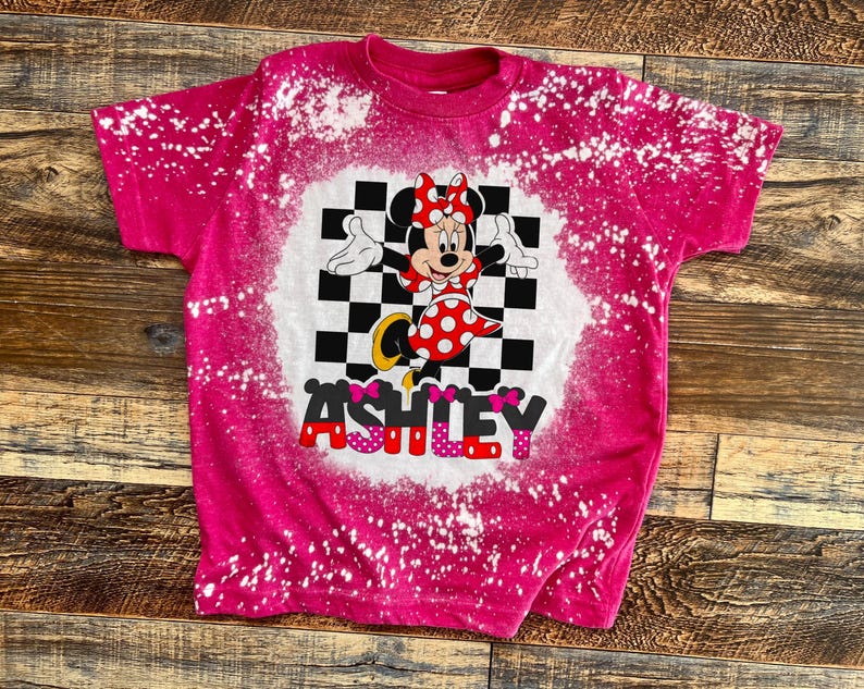 May include: Pink t-shirt with a bleach-splattered design featuring a cartoon image of Minnie Mouse in a checkered background with the name "ASHLEY" in pink letters.