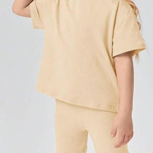 May include: A two-piece outfit for kids in a light beige color. The outfit includes a short-sleeved t-shirt and matching bike shorts.