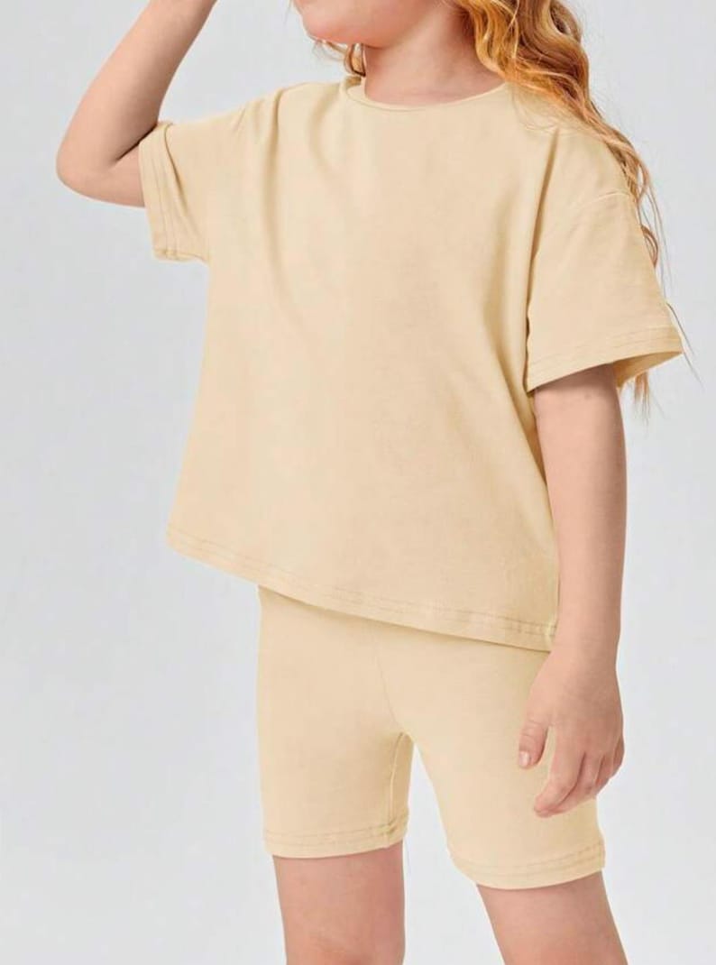 May include: A two-piece outfit for kids in a light beige color. The outfit includes a short-sleeved t-shirt and matching bike shorts.