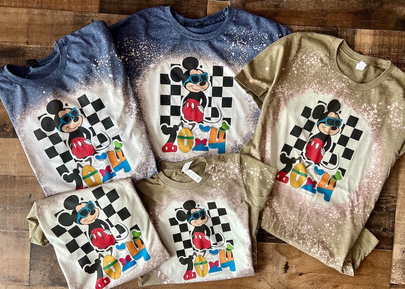 May include: Four bleached t-shirts with a cartoon image of Mickey Mouse wearing sunglasses and standing in front of a checkered pattern. The text "2024" is below Mickey Mouse. The shirts are in different colors: blue, green, white, and white.