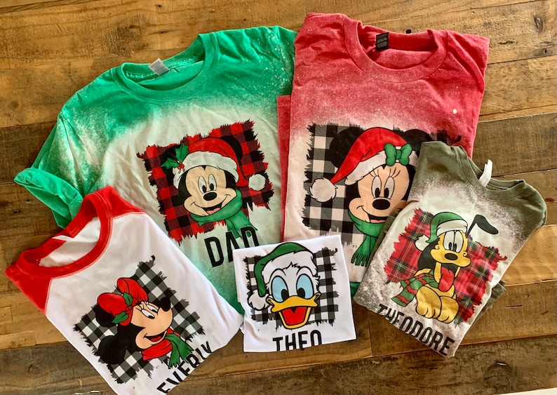 May include: Five green, red, and white t-shirts with a bleach-splattered effect. Each shirt features a different Disney character wearing a Santa hat and a plaid scarf. The shirts are labeled "Dad", "Everly", "Theo", "Theodore", and "Mom".