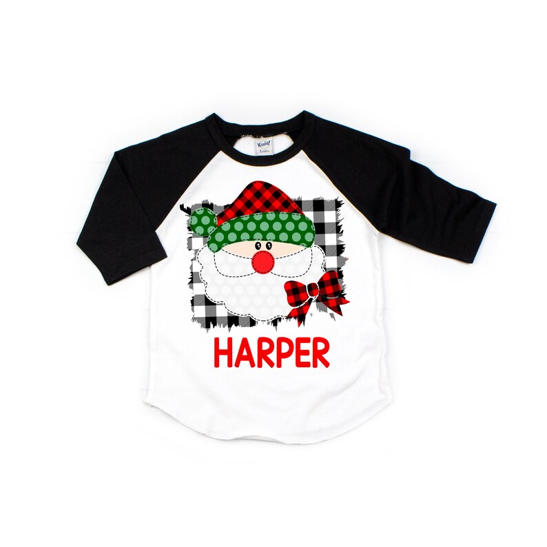May include: A white, black, and red baseball tee shirt with a Santa Claus face graphic. The Santa Claus graphic has a red and black plaid hat, a green hat with white polka dots, a white beard, and a red bow. The shirt has the name "HARPER" printed in red letters below the graphic.