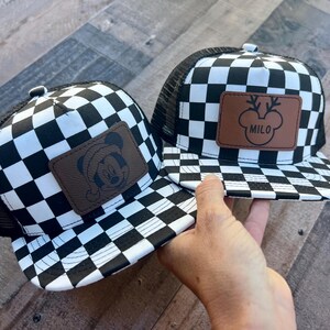 K&ouml;nnte beinhalten: Zwei schwarz-wei&szlig; karierte Trucker-H&uuml;te. Ein Hut hat einen braunen Lederaufn&auml;her mit einem Mickey Mouse-Design, das einen Weihnachtsmannhut tr&auml;gt. Der andere Hut hat einen braunen Lederaufn&auml;her mit einem Rentier-Design und dem Namen "MILO".