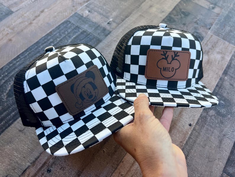 K&ouml;nnte beinhalten: Zwei schwarz-wei&szlig; karierte Trucker-H&uuml;te. Ein Hut hat einen braunen Lederaufn&auml;her mit einem Mickey Mouse-Design, das einen Weihnachtsmannhut tr&auml;gt. Der andere Hut hat einen braunen Lederaufn&auml;her mit einem Rentier-Design und dem Namen "MILO".