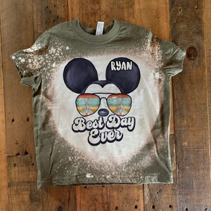 May include: A green t-shirt with a bleach-splattered design featuring a cartoon Mickey Mouse wearing sunglasses with the words "Best Day Ever" and the name "Ryan" on the shirt.