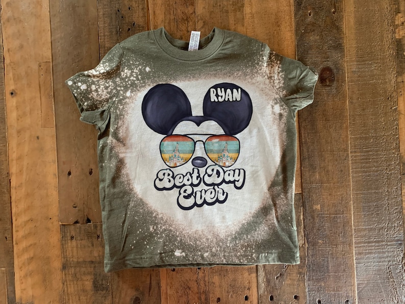 May include: A green t-shirt with a bleach-splattered design featuring a cartoon Mickey Mouse wearing sunglasses with the words "Best Day Ever" and the name "Ryan" on the shirt.