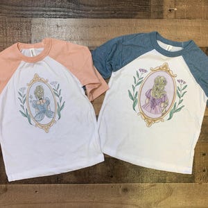 Peut inclure: Deux t-shirts de baseball blancs et p&ecirc;che avec un motif de princesse dans un cadre dor&eacute;. La princesse porte une robe bleue et a les cheveux blonds. Les t-shirts ont un design bicolore bleu et p&ecirc;che.