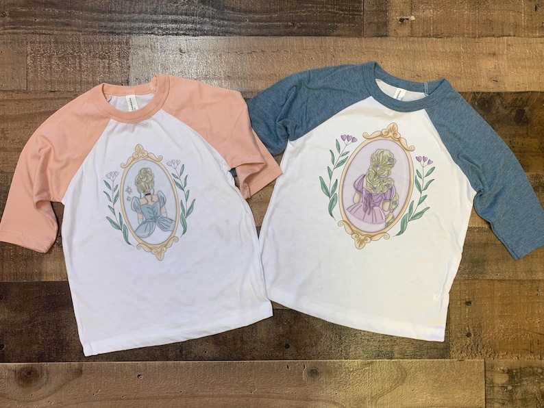 Peut inclure: Deux t-shirts de baseball blancs et p&ecirc;che avec un motif de princesse dans un cadre dor&eacute;. La princesse porte une robe bleue et a les cheveux blonds. Les t-shirts ont un design bicolore bleu et p&ecirc;che.