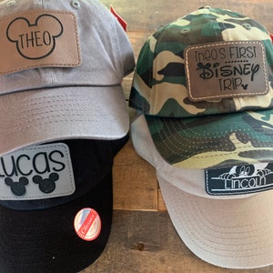 May include: Four baseball caps in grey, black, camouflage, and beige. Each cap has a leather patch with a name or text, including "THEO", "LUCAS", "Theo's FIRST DISNEY TRIP", and "LINCOLN".