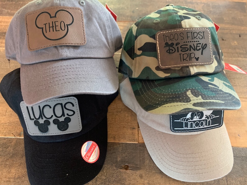 May include: Four baseball caps in grey, black, camouflage, and beige. Each cap has a leather patch with a name or text, including "THEO", "LUCAS", "Theo's FIRST DISNEY TRIP", and "LINCOLN".