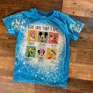 May include: A blue t-shirt with a bleach-splattered design featuring a graphic of Disney characters with bible verses. The text reads "God says that I am..." with each character representing a different verse.