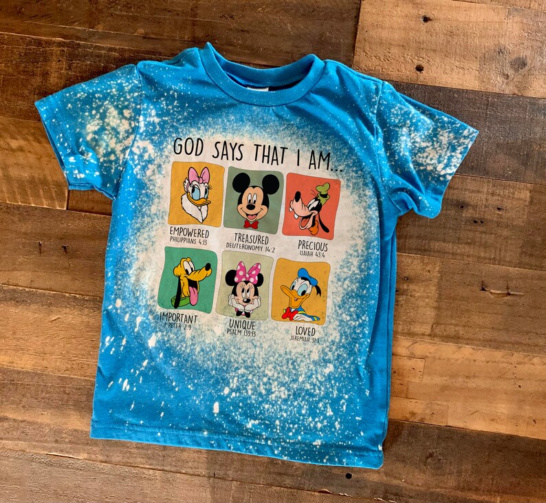 May include: A blue t-shirt with a bleach-splattered design featuring a graphic of Disney characters with bible verses. The text reads "God says that I am..." with each character representing a different verse.