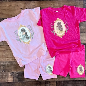 Peut inclure: Deux t-shirts &agrave; manches courtes, l'un rose et l'autre fuchsia, avec des shorts assortis. Le t-shirt rose pr&eacute;sente une illustration encadr&eacute;e de deux femmes avec les cheveux en chignon. Le t-shirt fuchsia pr&eacute;sente une illustration encadr&eacute;e d'une femme aux cheveux longs tenant une rose.