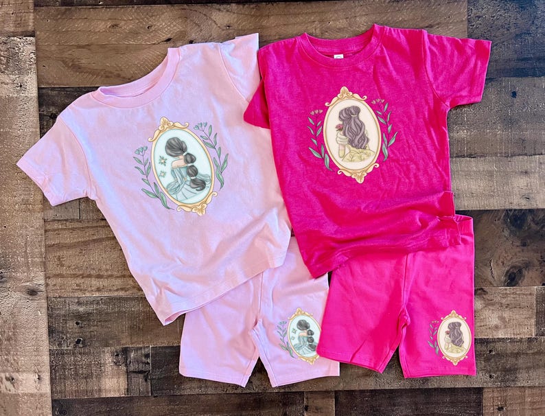 Peut inclure: Deux t-shirts &agrave; manches courtes, l'un rose et l'autre fuchsia, avec des shorts assortis. Le t-shirt rose pr&eacute;sente une illustration encadr&eacute;e de deux femmes avec les cheveux en chignon. Le t-shirt fuchsia pr&eacute;sente une illustration encadr&eacute;e d'une femme aux cheveux longs tenant une rose.