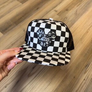 May include: A black and white chequered trucker hat with a mesh back. The front has a Mickey Mouse-shaped patch with Star Wars characters and text. The hat has a curved peak and a button on top.