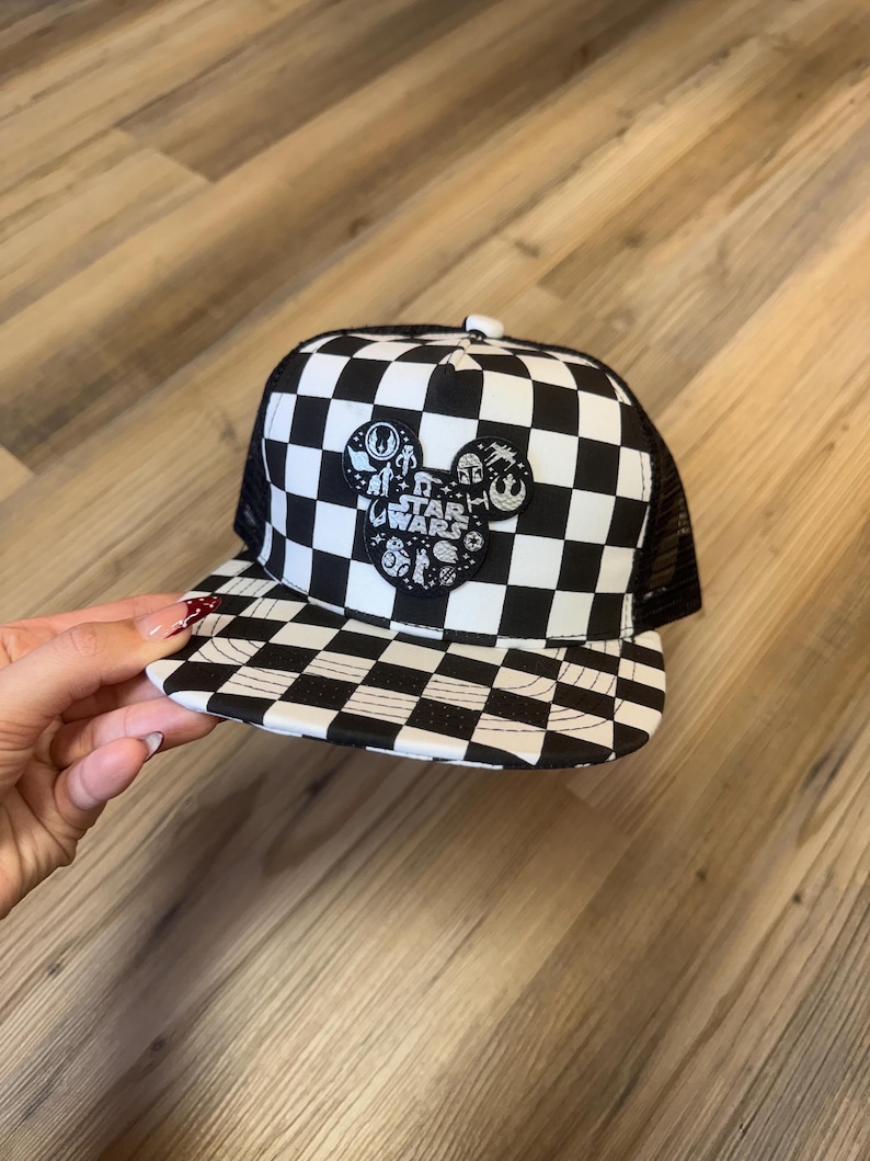 May include: A black and white chequered trucker hat with a mesh back. The front has a Mickey Mouse-shaped patch with Star Wars characters and text. The hat has a curved peak and a button on top.