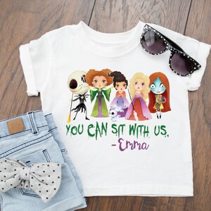 Puede incluir: Camiseta blanca con un gr&aacute;fico verde, morado y negro de personajes de dibujos animados de las pel&iacute;culas "Hocus Pocus" y "Pesadilla antes de Navidad". El texto "You can sit with us. -Emma" est&aacute; impreso debajo del gr&aacute;fico.