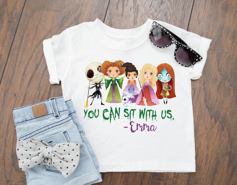 Puede incluir: Camiseta blanca con un gr&aacute;fico verde, morado y negro de personajes de dibujos animados de las pel&iacute;culas "Hocus Pocus" y "Pesadilla antes de Navidad". El texto "You can sit with us. -Emma" est&aacute; impreso debajo del gr&aacute;fico.