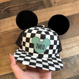 May include: A black and white chequered baseball cap with black mesh backing and large black circular ears. A light green patch with the name "THEO" is centered on the front of the cap.