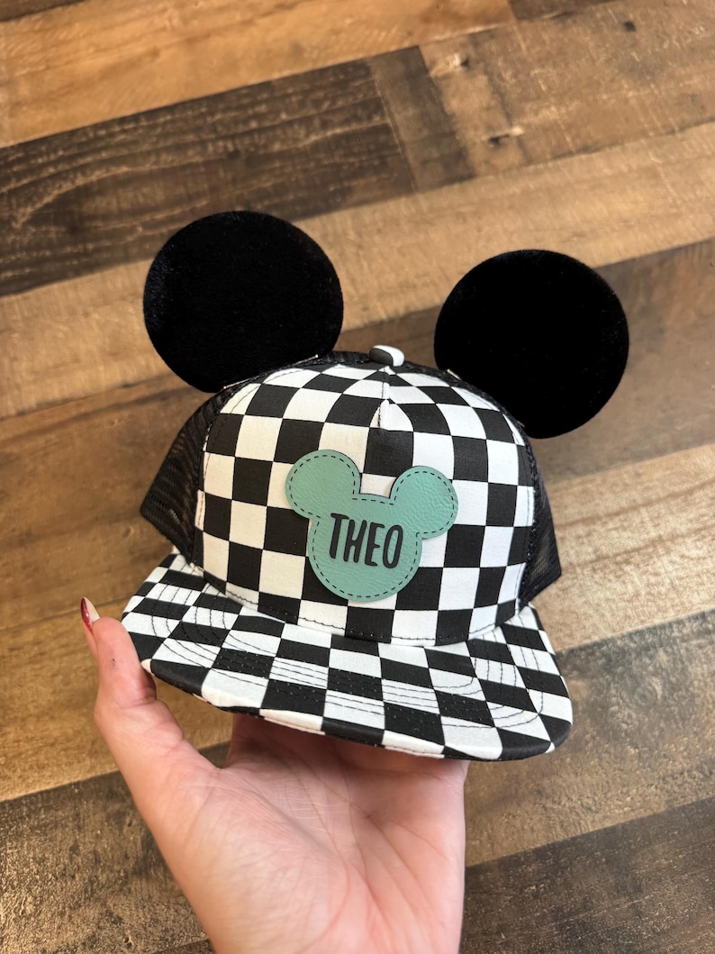 May include: A black and white chequered baseball cap with black mesh backing and large black circular ears. A light green patch with the name "THEO" is centered on the front of the cap.