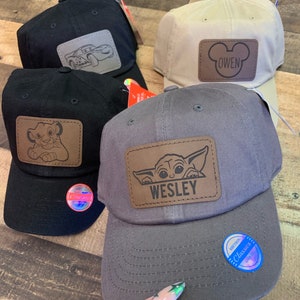May include: Four baseball caps in black, beige, and grey. Each cap features a brown leather patch with a cartoon design or name. Designs include a car, Simba, Mickey Mouse with the name "Owen", and Baby Yoda with the name "Wesley".