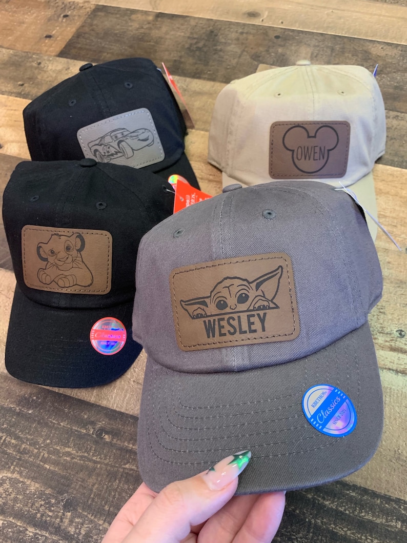 May include: Four baseball caps in black, beige, and grey. Each cap features a brown leather patch with a cartoon design or name. Designs include a car, Simba, Mickey Mouse with the name "Owen", and Baby Yoda with the name "Wesley".