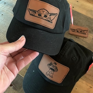 May include: Black baseball caps with brown leather patches. One cap features a cartoon image of Grogu, and the other features Buzz Lightyear. The caps have a curved peak and a tag.