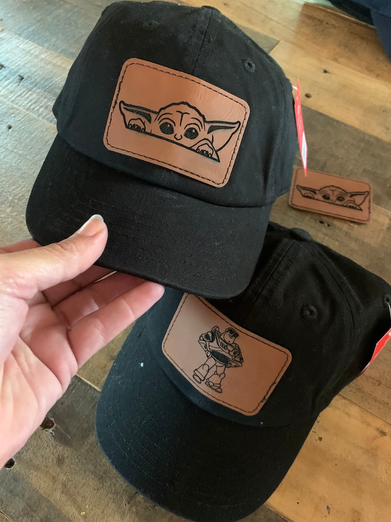 May include: Black baseball caps with brown leather patches. One cap features a cartoon image of Grogu, and the other features Buzz Lightyear. The caps have a curved peak and a tag.