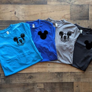 May include: Four short-sleeved t-shirts in blue, royal blue, gray, and black. Each shirt features a Mickey Mouse graphic. The blue shirt has a sketched Mickey face, the royal blue and black shirts have a Mickey head silhouette.