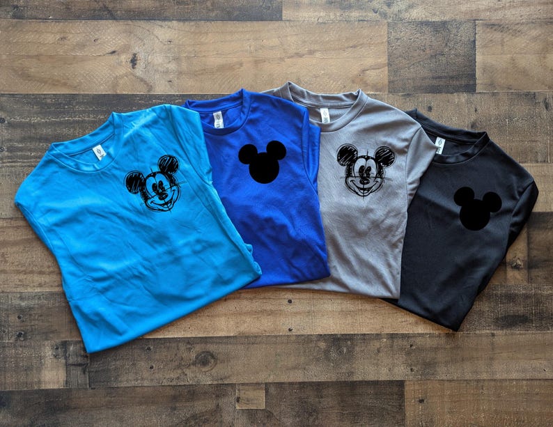 May include: Four short-sleeved t-shirts in blue, royal blue, gray, and black. Each shirt features a Mickey Mouse graphic. The blue shirt has a sketched Mickey face, the royal blue and black shirts have a Mickey head silhouette.