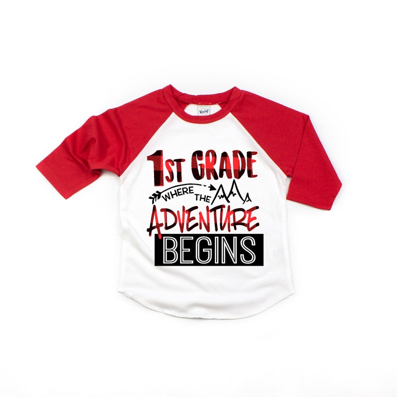 May include: A white and red baseball tee shirt with a black and red plaid design. The shirt has the text "1st Grade Where the Adventure Begins" printed on the front.