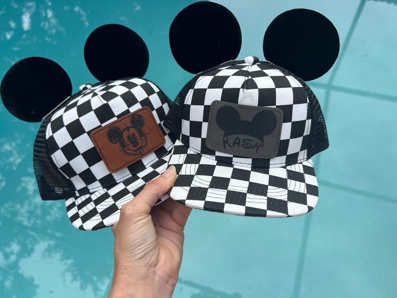 K&ouml;nnte beinhalten: Zwei schwarz-wei&szlig; karierte Baseballkappen mit schwarzen Mesh-R&uuml;cken und gro&szlig;en schwarzen Micky-Maus-Ohren. Eine Kappe hat einen braunen Lederaufn&auml;her mit Mickey Mouse, die andere einen grauen Aufn&auml;her mit dem Namen "KASY".
