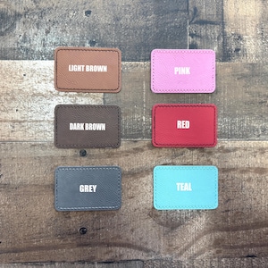 May include: Six rectangular leather patches in various colours: light brown, dark brown, pink, red, grey, and teal. Each patch has a white text label indicating its colour. The patches are arranged on a wooden surface.