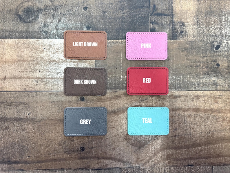 May include: Six rectangular leather patches in various colours: light brown, dark brown, pink, red, grey, and teal. Each patch has a white text label indicating its colour. The patches are arranged on a wooden surface.