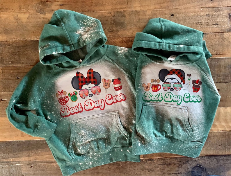 May include: Two green hooded sweatshirts with a bleach-splattered design. The sweatshirts feature a Christmas-themed graphic with Mickey Mouse and friends, along with the text "Best Day Ever".