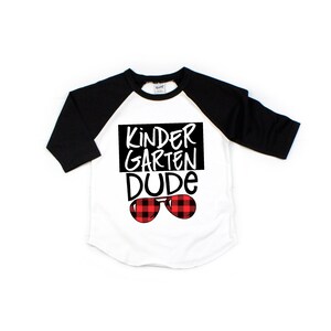 May include: White and black baseball tee shirt with red and black plaid sunglasses and the text "Kindergarten Dude" in black.