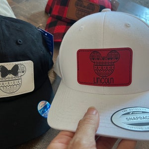 May include: Two black and white embroidered Mickey Mouse patches on baseball caps. One cap is black and the other is white. The white cap has a red patch with the name "Lincoln" on it. The caps have a "The Authentic Yupoong Snapback" label.