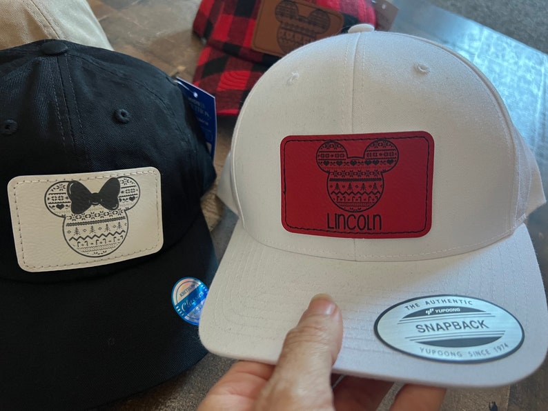 May include: Two black and white embroidered Mickey Mouse patches on baseball caps. One cap is black and the other is white. The white cap has a red patch with the name "Lincoln" on it. The caps have a "The Authentic Yupoong Snapback" label.