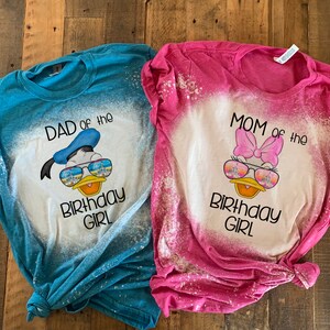 May include: Two bleached t-shirts with a Disney character design. The blue shirt says "Dad of the Birthday Girl" and features a cartoon Donald Duck wearing sunglasses. The pink shirt says "Mom of the Birthday Girl" and features a cartoon Daisy Duck wearing sunglasses.