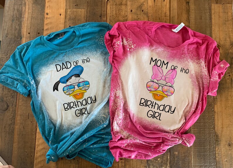 May include: Two bleached t-shirts with a Disney character design. The blue shirt says "Dad of the Birthday Girl" and features a cartoon Donald Duck wearing sunglasses. The pink shirt says "Mom of the Birthday Girl" and features a cartoon Daisy Duck wearing sunglasses.