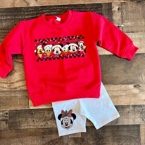 Peut inclure: Un sweat-shirt rouge avec un motif de personnages Disney portant des bonnets de Père Noël et des guirlandes lumineuses. Il est associé à un short blanc avec un motif de Minnie. Ensemble pour enfants.