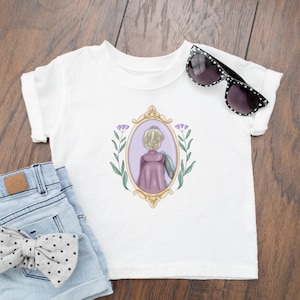 Peut inclure: T-shirt blanc avec une illustration violette et dor&eacute;e d'une femme aux cheveux blonds dans un cadre avec des fleurs violettes. La femme porte une cape violette et regarde loin du spectateur.